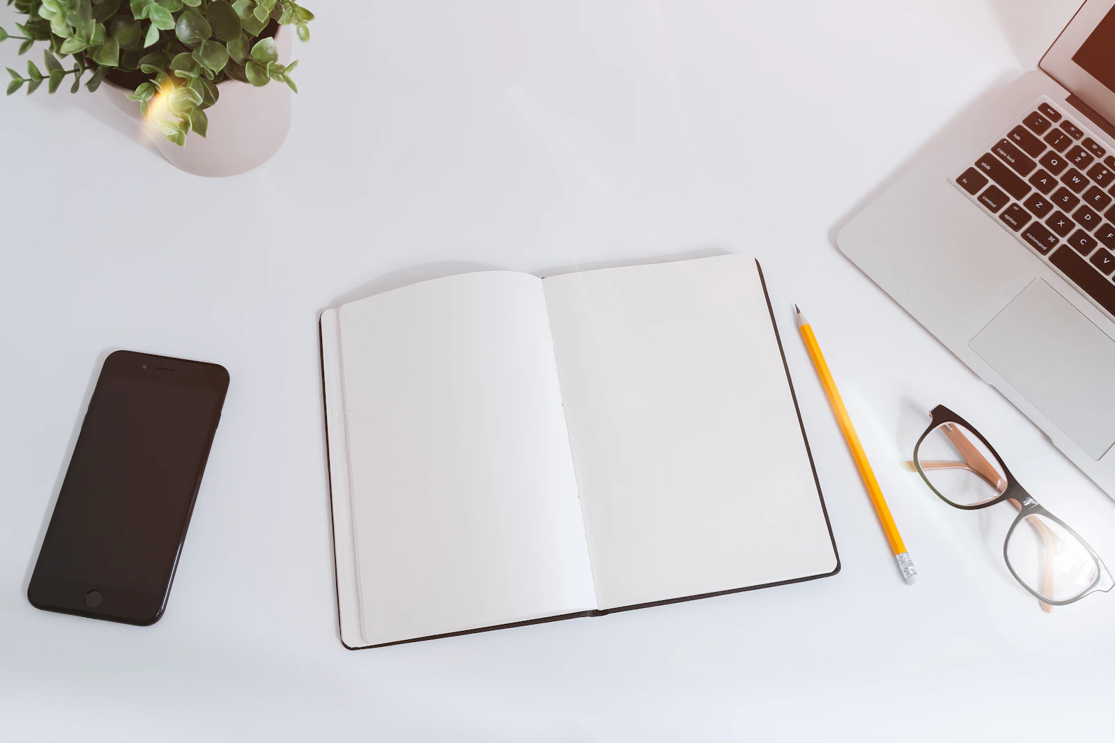 Caderno aberto ao lado de um computador, um celular e óculos